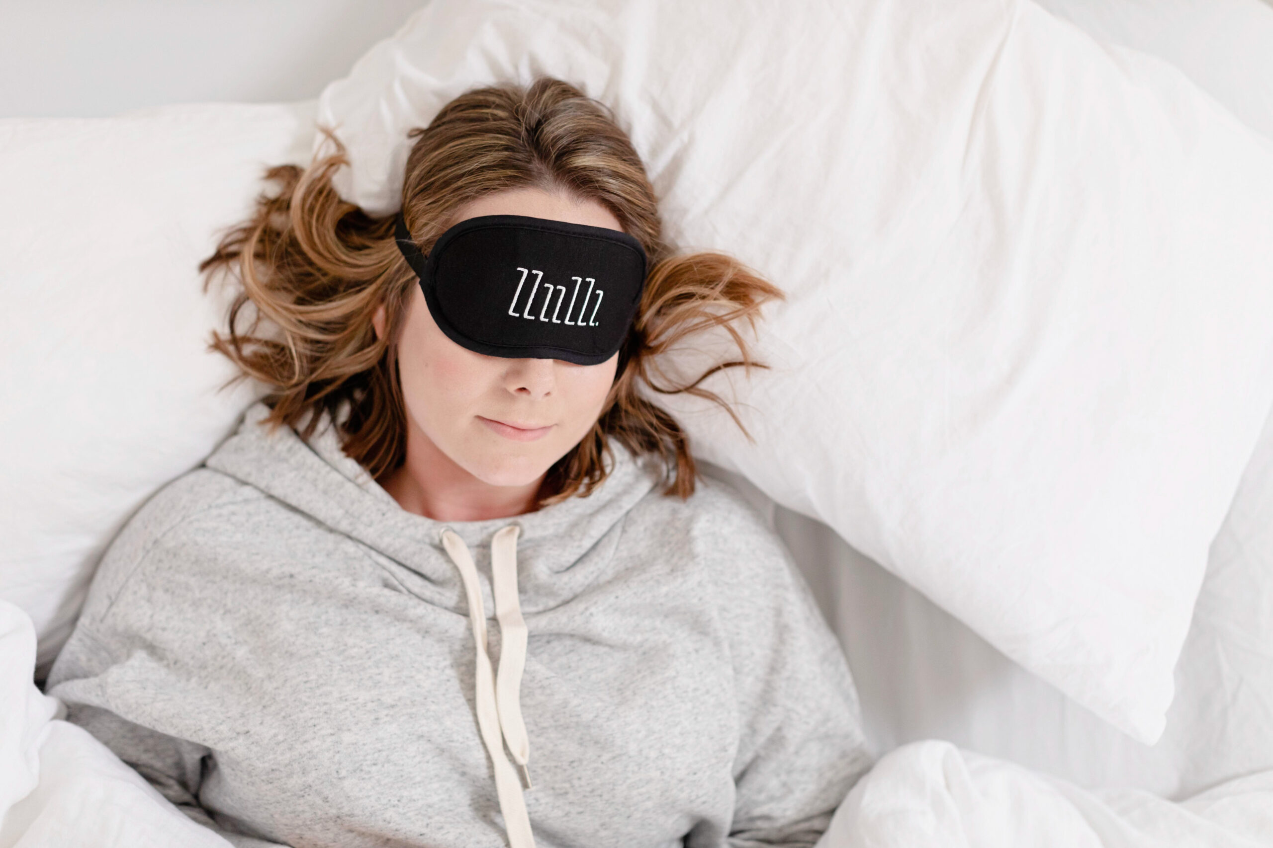 woman wearing gray sweatshirt and black sleep mask with zzzz's laying on white pillow