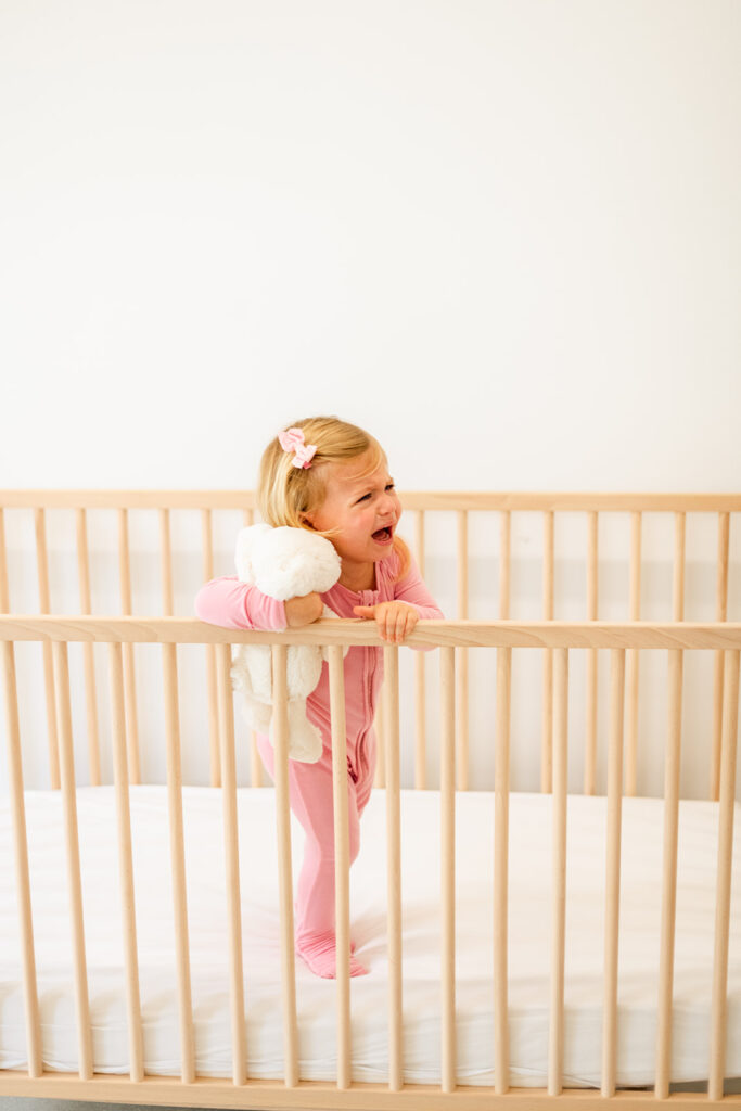 A two year old child with blond hair, wearing pink pajamas and holding a white stuffed animal, who is standing up in a crib crying. 